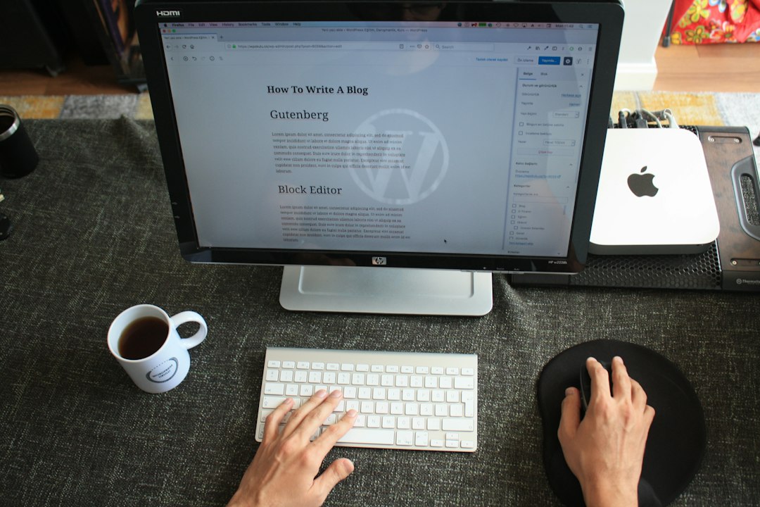 O persoană tastează pe o tastatură albă la un birou, editând o postare despre cum să scrii un articol de blog despre găzduirea WordPress pe un monitor mare. O ceașcă de cafea, un mousepad și accesorii tehnologice sunt aranjate ordonat în apropiere.